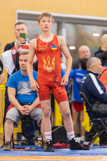 Young Ukrainian wrestler stands alert at ringside, wearing red singlet with trident emblem, composed before his match.