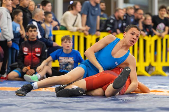 Teenage wrestler in blue singlet pins opponent in red on the mat, expression focused and intense, crowd watching behind.