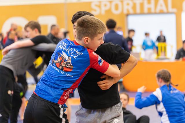 Young wrestler in blue PSK Olymp Praha jersey clinches opponent during warm-up, focused and determined expression.