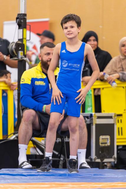 Young boy in blue Adidas singlet stands confidently on the mat, hands on thighs, coach seated behind him.