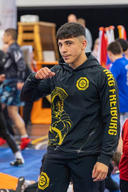 Teenage wrestler in black KKG Freiburg hoodie strikes a confident pose, fist raised, at a busy sports hall.