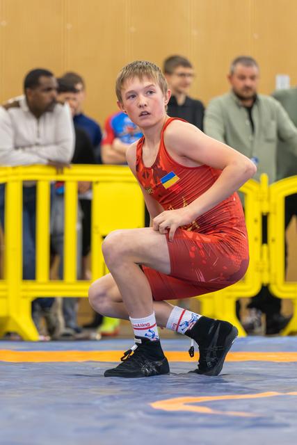 Young Ukrainian wrestler in red singlet crouches in a focused, ready stance on the blue mat during a match.