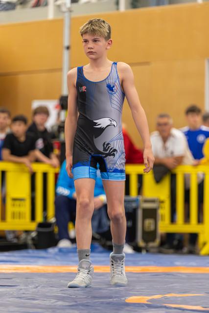 Young boy in eagle-design singlet walks confidently across the mat, composed expression, indoor wrestling venue in background.