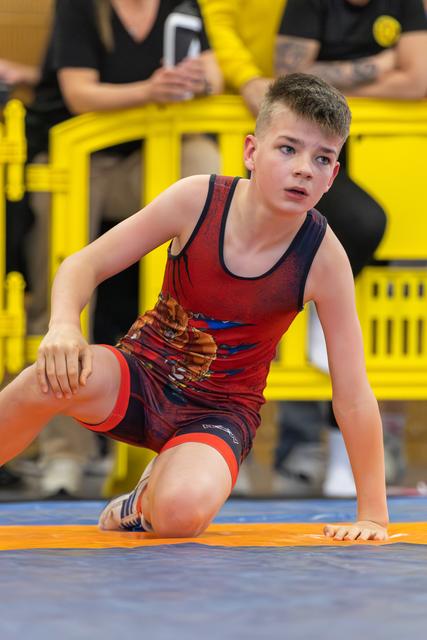 Focused young boy in red singlet kneels on the mat, alert and ready, with spectators seated behind yellow barriers.