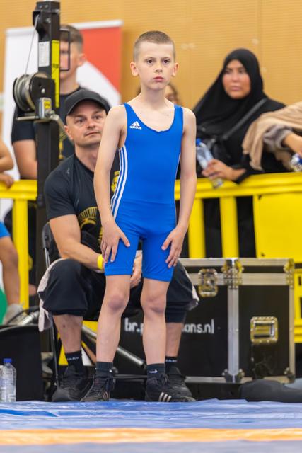Young boy in blue Adidas singlet stands poised on the mat, composed and focused, coach crouching attentively behind him.