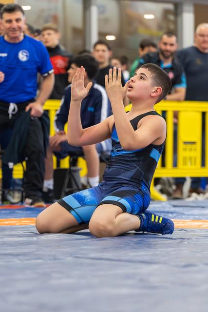 Young wrestler on his knees, arms raised in celebration, looking upward with joy after winning his match.
