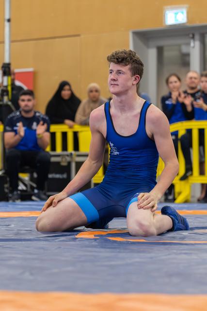 Young male wrestler in blue singlet kneels on the mat, gazing upward with quiet anticipation as spectators applaud behind him.