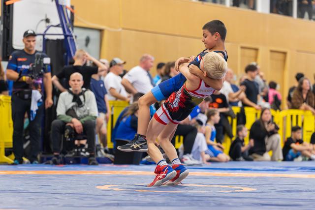 Blond boy lifts his opponent off the mat in a powerful wrestling move during a youth sports event.