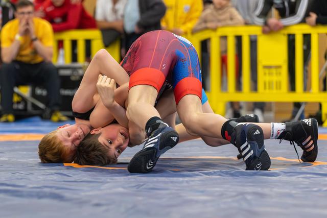 Two young wrestlers grapple on the mat, one dominant on top controlling the other in a tight hold, both straining with effort.