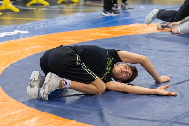 Young wrestler pinned on blue mat, straining to resist, face showing intense effort during a match.