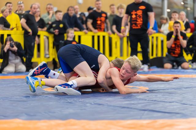 A young boy in red singlet is pinned to the mat by his opponent in blue during a youth wrestling match, crowd watching.