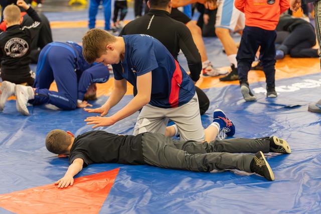 A boy in a blue shirt pins his opponent face-down on the mat, pressing firmly on his back during a match.