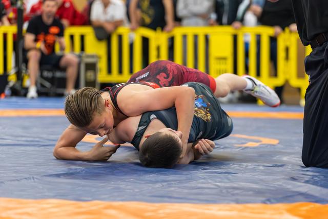 Young boy in red singlet pins his opponent to the mat, focused and determined, at an indoor youth wrestling event.