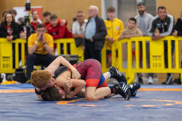 Young redhead wrestler pins opponent to the mat while spectators watch intently from behind yellow barriers.