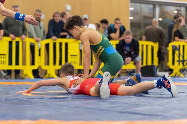 Boy in green singlet dominates opponent in red, pinning him to the blue mat during a youth wrestling bout.