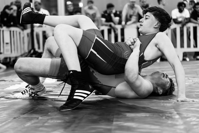 Young wrestler in dark singlet pins opponent to the mat, gripping firmly with focused determination during a match.