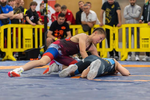 Young wrestler in red singlet dominates opponent pinned to the mat, crowd watching intently behind yellow barriers.