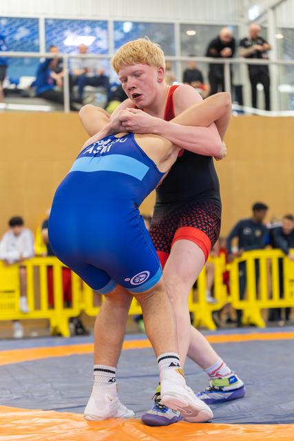 Blond teen wrestler in red singlet locks arms around opponent in blue, straining forward with intense focus on orange mat.