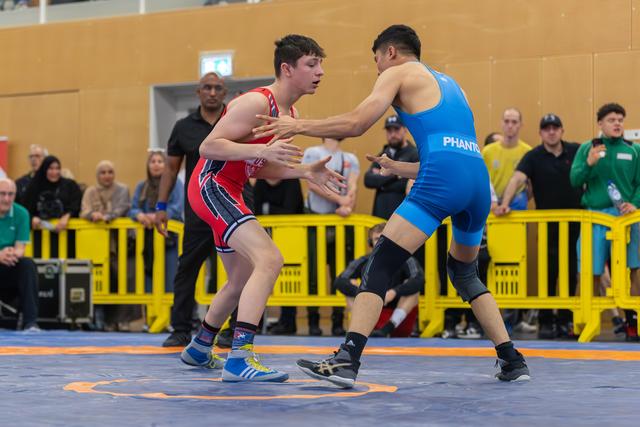 Teen wrestler in red singlet squares off against opponent in blue Phantom singlet on a competition mat, crowd watching.