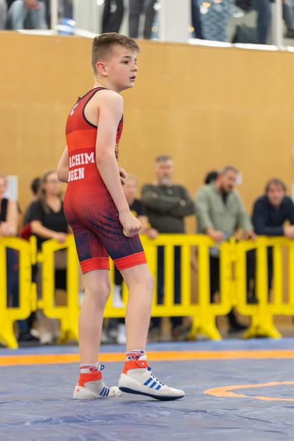 Young boy in red singlet stands alert on the mat, fists clenched, focused and ready during a youth wrestling event.