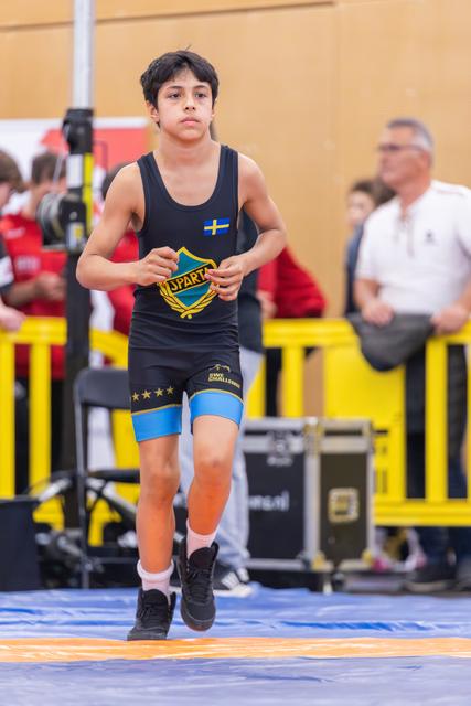 Focused young wrestler in black Sparta singlet with Swedish flag patch jogging onto the mat at an indoor sports event.