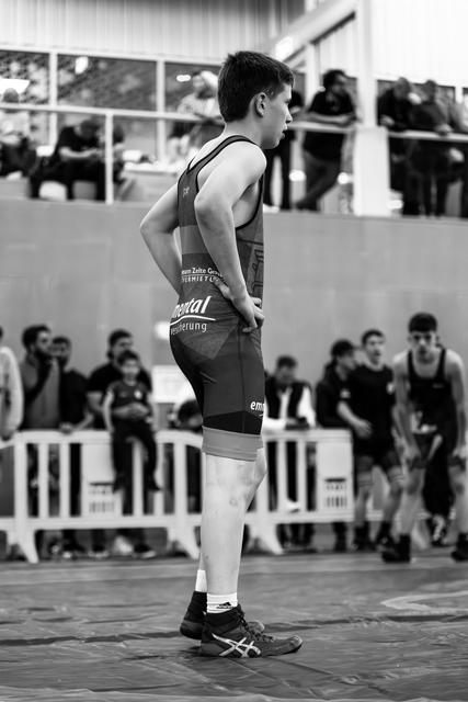 Teen wrestler stands still, hands on hips, composed and focused, watching match from the side of the mat.