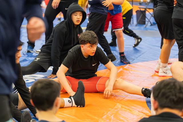 Young male wrestler in red shorts sits on orange mat stretching, looking focused and alert amid teammates warming up.