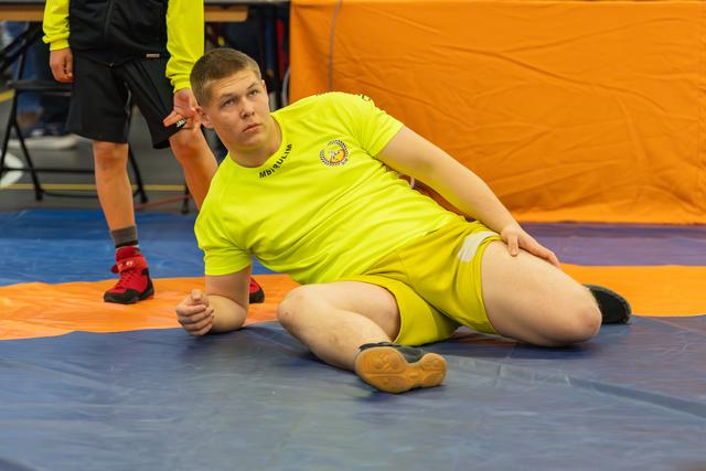 Young wrestler in neon yellow kit stretches on the mat, looking focused and composed during warm-up.
