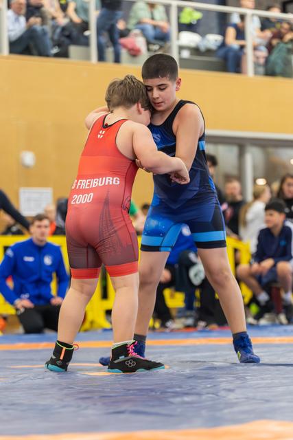 Two youth wrestlers engage in a standing clinch on the mat, one in red Freiburg 2000 singlet, the other in blue.