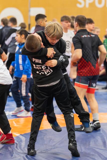 Two boys in black warm-up suits grapple on a blue mat, one lifting the other during warm-up at a busy sports hall.