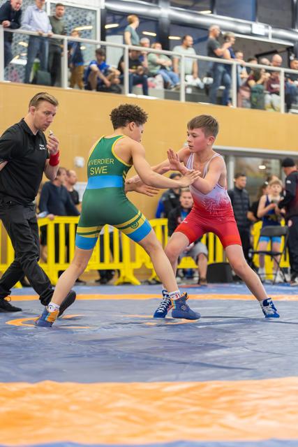 Two young wrestlers in green and red singlets square off on the mat, referee watching closely in an indoor arena.