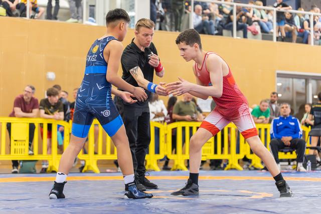 Two young wrestlers in blue and red singlets square off tensely as a referee watches closely in a busy arena.