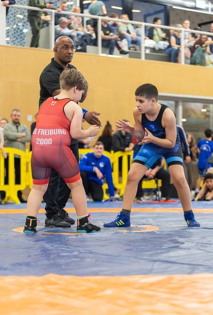 Two youth wrestlers in red and blue singlets square off on the mat, referee standing between them in a sports hall.