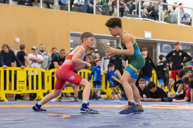 Two youth wrestlers in red and green singlets face off in athletic stance, crowd watching in background.