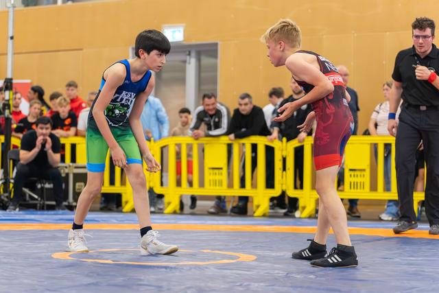 Two young wrestlers in blue and red singlets square off on the mat, focused and ready to engage, referee watching nearby.