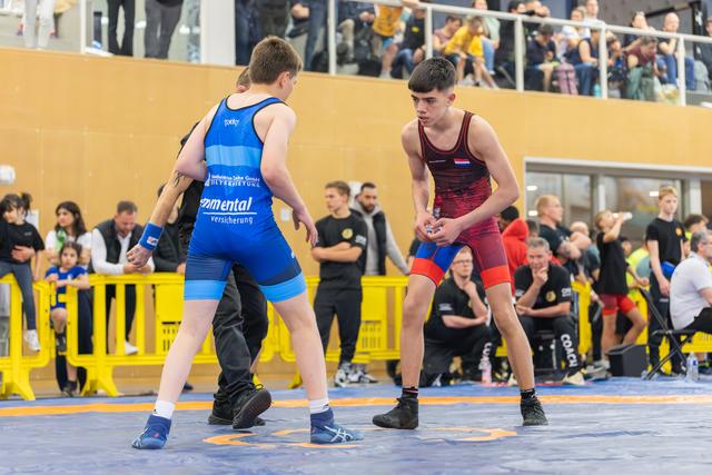 Two teen wrestlers in blue and red singlets square off on the mat, focused and ready to engage at an indoor event.