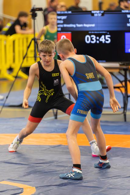 Two boys in wrestling stances face each other on the mat, focused and alert. Timer shows 3:45 remaining.