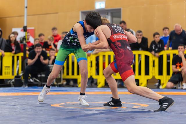 Two young wrestlers lock grips in intense clinch on blue mat, watched by crowd behind yellow barriers.
