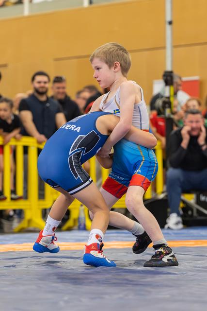 Two young boys locked in an intense wrestling grip on a blue mat, spectators watching closely in the background.
