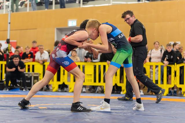 Two young wrestlers lock grips in intense focus on a blue competition mat, referee watching closely behind.
