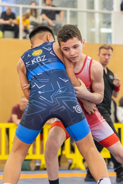 Two young wrestlers locked in a tight clinch, the red-suited athlete gripping his KGZ opponent with focused determination.