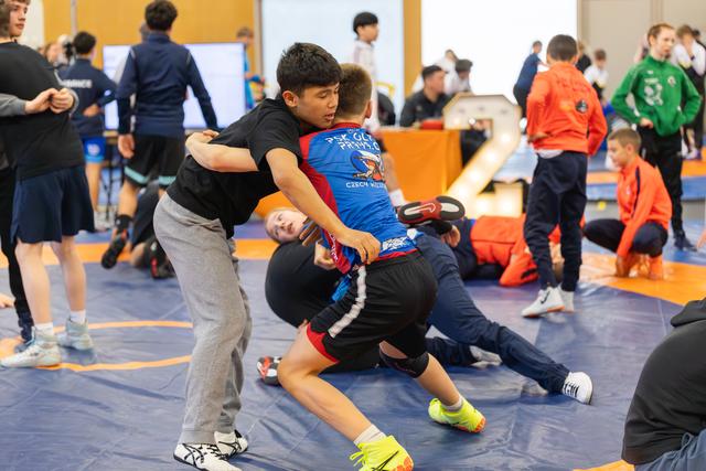 Two boys locked in an intense grapple on a blue mat, surrounded by other young wrestlers practicing in a busy hall.