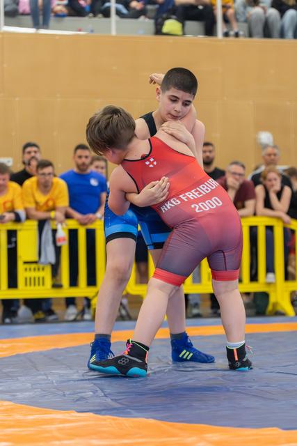 Two young wrestlers locked in a fierce clinch on the mat, red singlet competitor from RKG Freiburg 2000 holding firm.