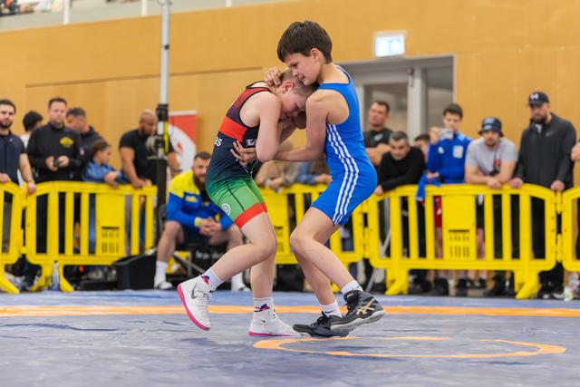 Two youth wrestlers locked in a tight clinch on the mat, both straining with effort during a competitive bout.