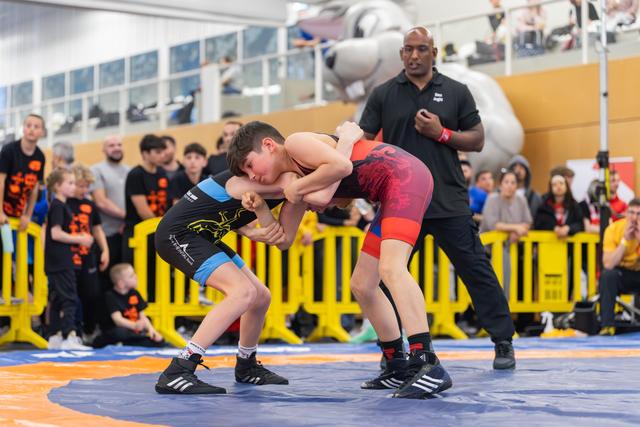 Two boys locked in an intense grapple on the mat, referee watching closely as spectators look on from behind barriers.