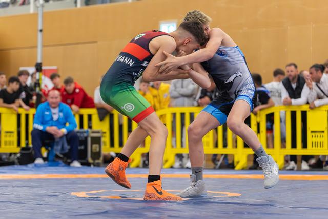 Two youth wrestlers lock in a fierce clinch on the blue mat, one lifted off his feet during an indoor competition.