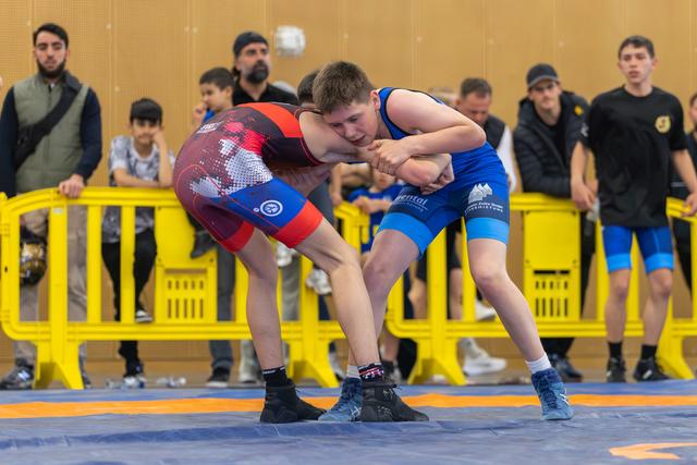 Two young wrestlers in a fierce clinch on the mat, spectators watching behind yellow barriers in a sports hall.