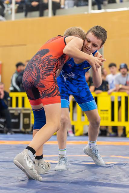 Two young wrestlers locked in a tight clinch on the mat, one in red, one in blue, both straining with focused determination.