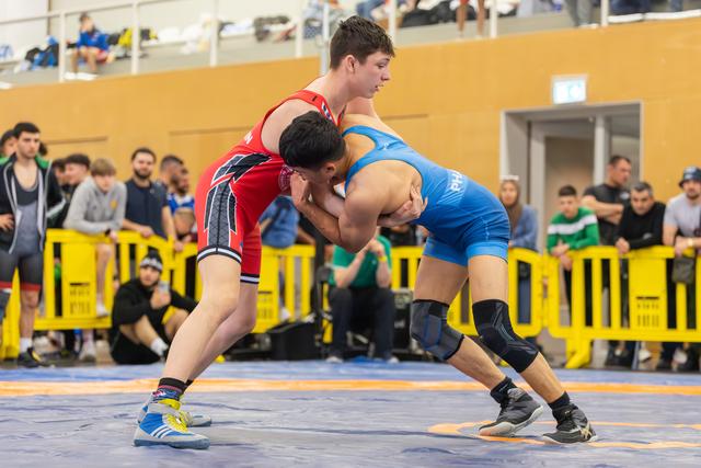 Two teen wrestlers lock in a tense upper-body clinch on the blue mat, spectators watching from behind yellow barriers.