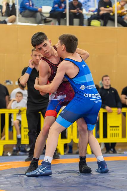 Two teen wrestlers locked in a grip battle, red singlet boy focused and determined, blue singlet opponent from team Emmental.